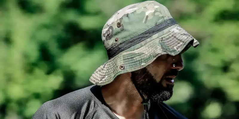 A man wearing a camouflage boonie hat with a chin strap, standing outdoors near water.