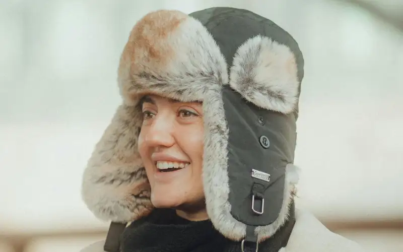 A woman wearing a dark charcoal winter trapper hat with faux fur ear flaps, smiling outdoors in cold weather