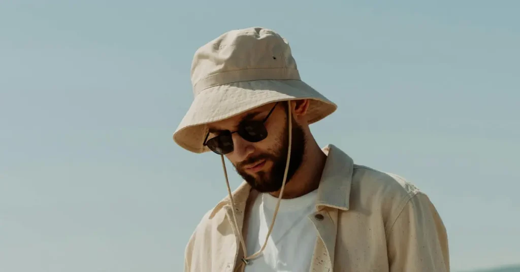 A man wearing a beige bucket hat with a chin strap, standing outdoors in sunlight, dressed in a casual cream shirt and sunglasses
