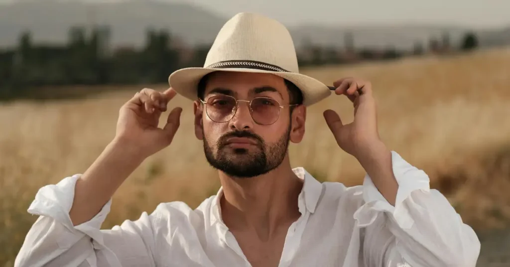 Man adjusting a light-colored fedora hat while wearing a white shirt in an outdoor field
