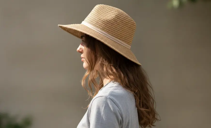 How to Wear a Hat with Long Hair Without Ruining Your Style 21 Side view of a woman with long wavy brown hair wearing a woven straw fedora hat with a white ribbon band, captured in natural outdoor lighting.