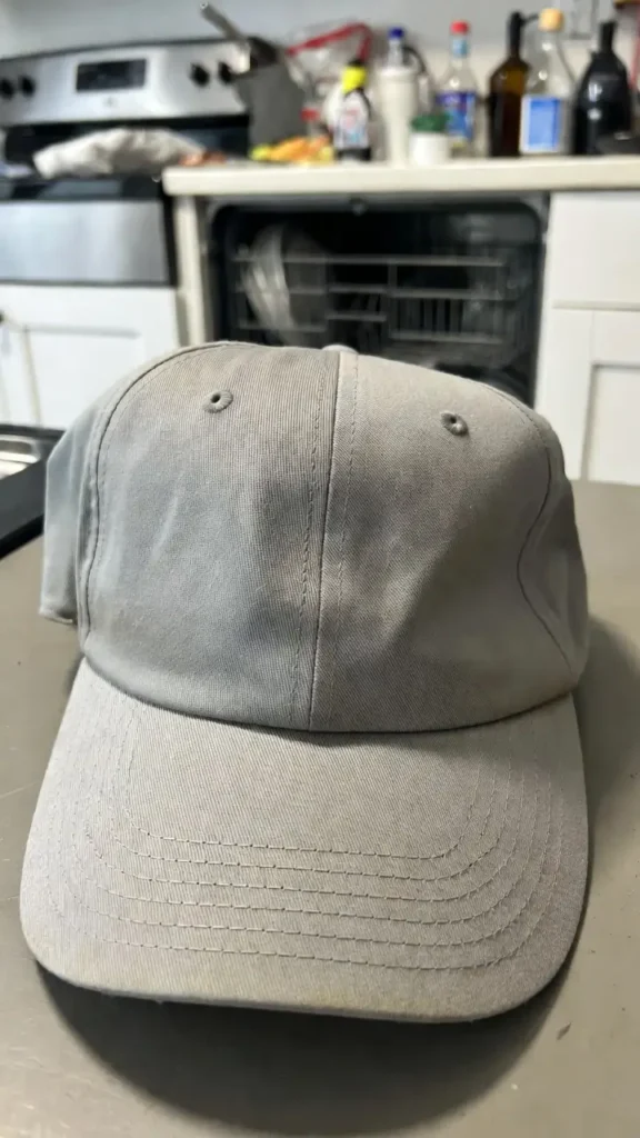 How to Wash a Hat Without Ruining It: Our Tested 2026 Guide 6 A grey baseball cap with a warped brim and faded fabric caused by high heat and harsh detergents in a dishwasher, shown on a kitchen counter.
