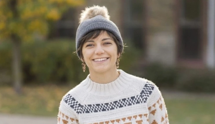 Woman with short hair wearing a grey pom-pom winter beanie and a patterned sweater outdoors.