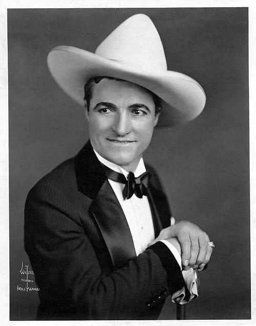 Studio portrait of silent film star Tom Mix wearing his signature tall cowboy hat and a tuxedo.