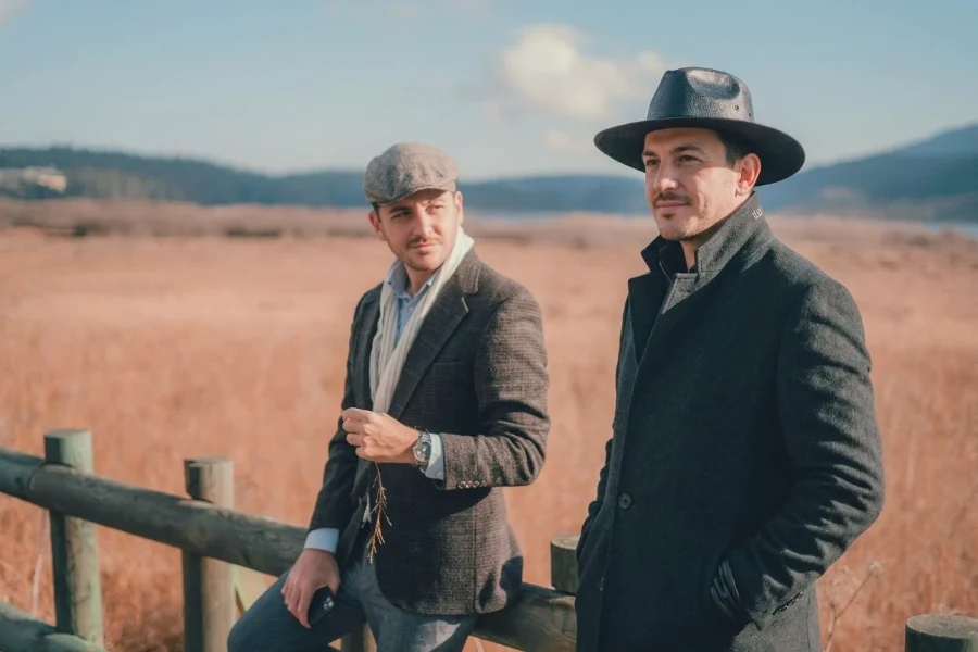 Home 17 Two men wearing a flat cap and a wide brim hat standing by a wooden fence in a field